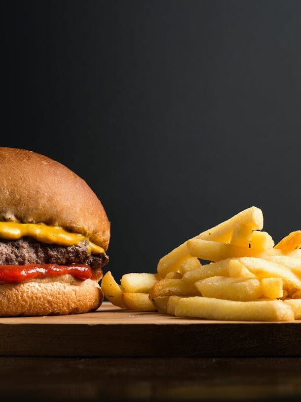 Appetizing burger with meat patty ketchup and cheese placed on wooden table with crispy french fries against black background